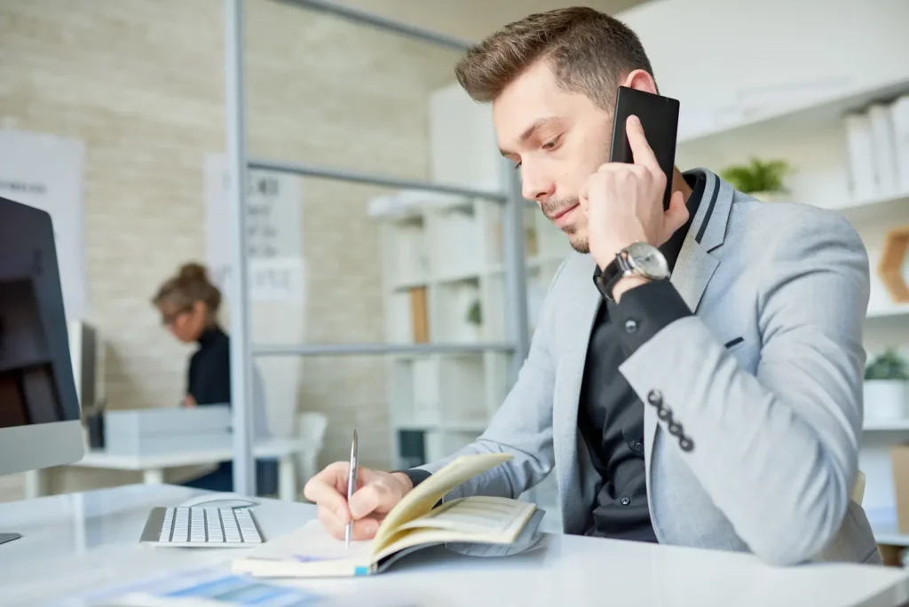 man-in-a-suit-talking-on-the-phone-while-writing-in-a-notebook-at-a-desk-with-a-computer-in-the-background-and-another-person-working