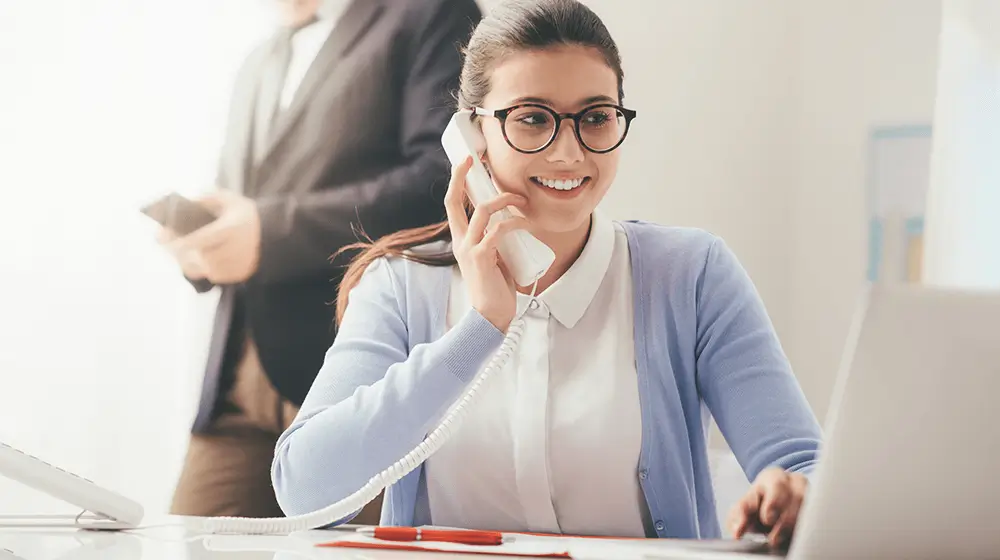 woman-with-glasses-smiling-and-talking-on-the-phone-while-sitting-at-a-desk-with-a-laptop-with-a-person-standing-in-the-background